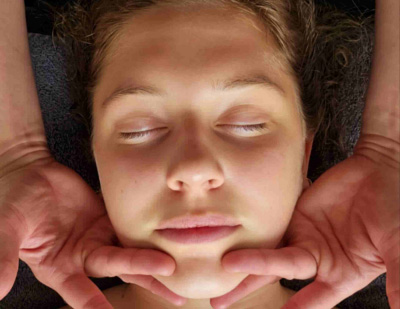 Image d’une jeune femme en train de recevoir un massage Kobido. Elle est couchée confortablement, les yeux fermés et elle paraît détendue. On voit les doigts de la praticienne qui sont en train de lisser la ligne de la mâchoire dans un geste qui part du menton vers les oreilles.