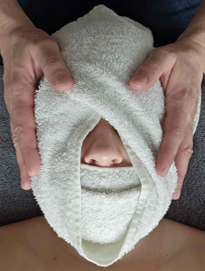 L’image montre une personne qui a son visage enveloppé par un oshibori chaud, les mains de la praticienne sont posées de chaque côté de l’oshibori. Seul le nez est à découvert et permet de bien respirer.